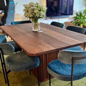 May include: A dark wood dining table with a rectangular top and a square base. The table is surrounded by six blue velvet chairs with black metal frames.
