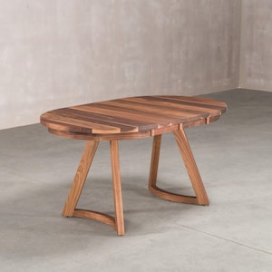May include: An oval dining table crafted from dark brown walnut wood with a visible grain pattern. The table features a smooth, rounded top and four angled, tapered legs that connect to curved base supports on each side of the furniture piece.