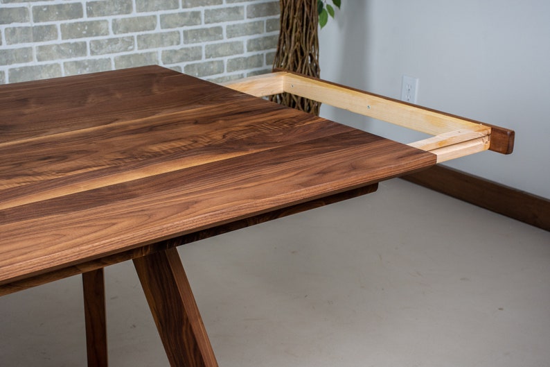 May include: A wooden dining table with a leaf extension. The table is made of walnut wood and has a natural finish. The leaf is made of maple wood and is attached to the table with a metal track.