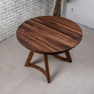 Puede incluir: Una mesa redonda de madera con un acabado en nogal marrón oscuro. La mesa tiene un diseño simple y moderno con una base curva.