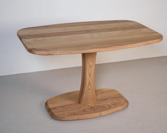 May include: A light-colored wooden table with a unique, organic design. The tabletop is oval-shaped, supported by a central, sculpted pedestal base. The table is made of light-colored wood, showcasing the natural grain.