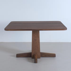 Peut inclure: Table carrée en bois de couleur marron foncé. Le plateau a des coins arrondis et repose sur un pied central avec quatre pieds. La table mesure environ 76 cm de long et de large.