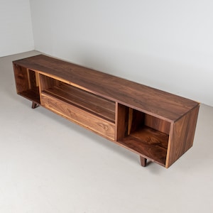 May include: A long, dark wood entertainment unit with a drawer and open shelves. The unit has a modern design with angled legs.