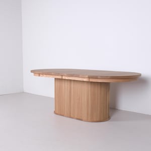 May include: A light brown, oval wooden table. The table features a solid, vertically-slatted base and a smooth tabletop. The design is modern and minimalist, suitable for a dining or work space. The table is approximately 180 cm long.