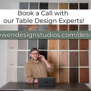 May include: A man in a brown shirt and glasses is on a phone call in front of a wall of wood samples. He is sitting at a table with a laptop in front of him. The text on the wall reads "Book a Call with our Table Design Experts!" and "loewendesignstudios.com/design".