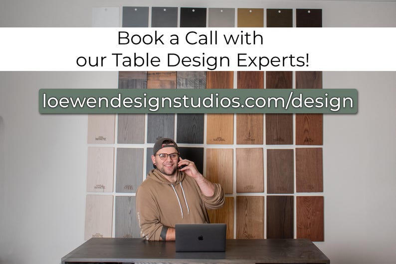 May include: A man in a brown shirt and glasses is on a phone call in front of a wall of wood samples. He is sitting at a table with a laptop in front of him. The text on the wall reads "Book a Call with our Table Design Experts!" and "loewendesignstudios.com/design".