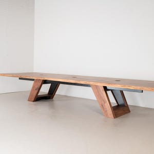 May include: A long rectangular table featuring a natural wood grain surface with a warm brown finish. The table is supported by two angled, trapezoidal legs constructed from a combination of matching wood and black metal structural supports.