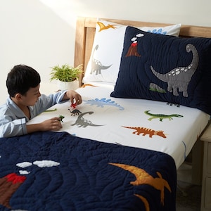 Puede incluir: Un juego de cama de temática de dinosaurios blanco y azul con un edredón acolchado azul marino. La ropa de cama presenta una variedad de dinosaurios, incluyendo un dinosaurio verde, un dinosaurio marrón y un dinosaurio gris. La ropa de cama también presenta un volcán y nubes.