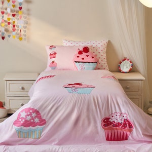 Puede incluir: Juego de cama rosa con temática de cupcakes con diseños de cupcakes bordados. El juego incluye un edredón y fundas de almohada, con varias ilustraciones de cupcakes con cerezas y estrellas. La paleta de colores es principalmente rosa, con acentos de azul, blanco y rojo.