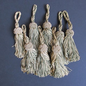 Peut inclure: Ensemble de huit glands décoratifs. Chaque gland est composé d'un nœud supérieur, d'une boucle en cordon torsadé et d'une longue frange. Les glands sont principalement beiges et verts, sur un fond neutre.