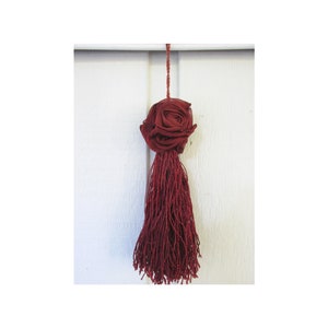 May include: A burgundy tassel with a rose-shaped top, hanging from a white rod. The rose is made of layered fabric, and the tassel is composed of numerous strands. The background is a white surface.