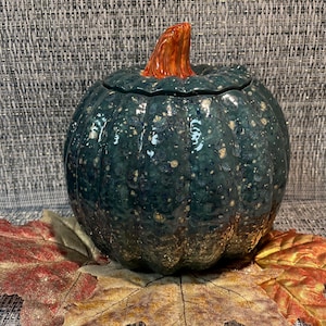 May include: A ceramic pumpkin-shaped container with a glossy, dark teal glaze featuring speckled gold accents. The lid is topped with a textured, orange-brown stem. The pumpkin rests on a base of several artificial autumn leaves in shades of red and gold.