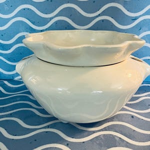 May include: A two-piece, off-white ceramic serving dish set. The bottom piece is a rounded bowl with two small handles. The top piece is a smaller, fluted dish. The set is placed on a blue and white wavy patterned surface.