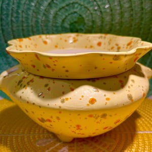 May include: A two-piece ceramic planter set. The base is a round, yellow pot with three small feet and two handles. A smaller, scalloped-edge dish sits on top. Both pieces are speckled with orange and brown dots. The set is on a yellow and green background.
