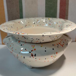 May include: A two-piece ceramic serving dish set. The white dishes have a speckled design with black, orange, and yellow dots. The bottom dish has handles and feet, while the top dish has a wide rim.