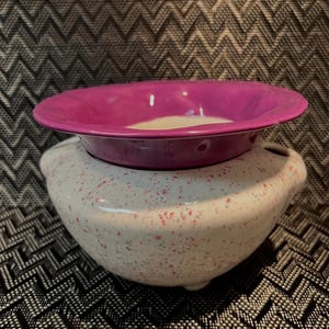 May include: A two-piece ceramic decorative bowl set. The top bowl is a vibrant magenta, while the base is white with pink speckles. The base has two small handles on either side. The set is designed for decorative purposes.