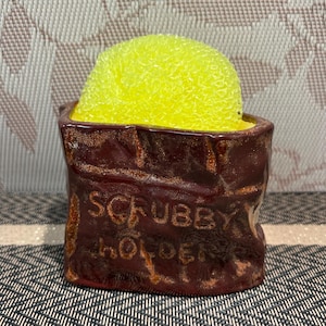 May include: A brown ceramic "Scrubby Holder" with a yellow sponge. The holder has a rustic, textured finish and the words "Scrubby Holder" are embossed on the front.