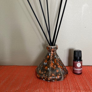 Peut inclure: Un diffuseur en céramique noir et orange avec des bâtonnets diffuseurs noirs et une petite bouteille d'huile essentielle de cannelle. La bouteille du diffuseur a un design géométrique et la bouteille d'huile essentielle a une étiquette brune avec le mot "Cannelle" imprimé dessus.