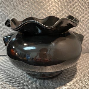 May include: A glossy black glass bowl featuring a wide, rounded body and a distinctively ruffled, wavy rim. The surface of the glass is smooth and reflective, showcasing a deep, solid black colour throughout the entire structure of the vessel.