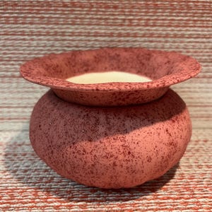 May include: A small, round ceramic bowl with a wide, flared rim. The exterior features a textured, speckled finish in shades of dusty rose and dark red. The interior of the bowl is smooth and off-white, contrasting with the mottled exterior surface.