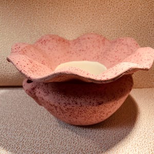 May include: A pink ceramic bowl with a scalloped, flower-like rim and a speckled texture throughout the exterior. The interior of the bowl is smooth and off-white, contrasting with the textured pink outer surface of the rounded vessel.