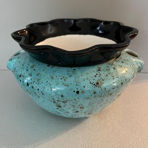 May include: A two-piece ceramic set. The bottom piece is a speckled turquoise bowl. The top piece is a black, scalloped-edge bowl. The interior of the top bowl is white. The set is likely for decorative or functional use.