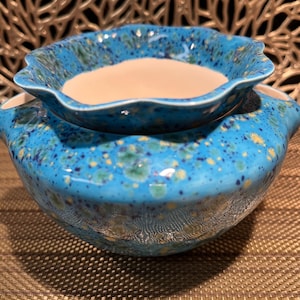 May include: A two-piece ceramic bowl set. The larger bowl is a vibrant turquoise with speckled accents of yellow, green, and dark blue. The smaller bowl, with a scalloped edge, sits atop the larger one. The interior of both bowls is a creamy beige.
