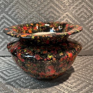 May include: A ceramic bowl with a wide, flared rim featuring a glossy black base covered in a vibrant, speckled pattern of red, orange, and yellow paint splatters. The bowl has a rounded body and a smooth, reflective finish throughout its surface.