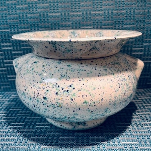 May include: A ceramic bowl with a wide, flared rim and two small side handles. The bowl features a cream-coloured base with a speckled pattern of dark blue, teal, and yellow dots across the entire exterior surface and the inner rim.