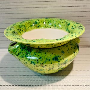 May include: A ceramic oil warmer featuring a bright lime green base with dark green and brown speckles. The top section is a wide, shallow bowl with a smooth white interior, designed to hold oil or wax, resting on a rounded, footed base.