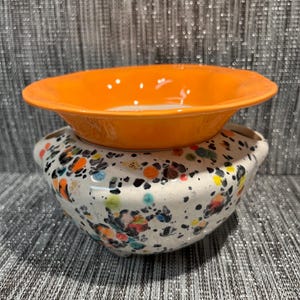 Peut inclure: Ensemble de bols en céramique. Le bol du bas est blanc avec des éclaboussures colorées. Le bol du haut est orange vif. Les bols sont empilés. Les bols ont une finition brillante.