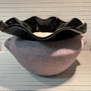 May include: A two-toned ceramic bowl featuring a textured, speckled lavender-grey base and a smooth, glossy black rim with a distinct wavy, ruffled edge. The interior of the bowl is white, contrasting with the dark exterior and the rough, granular surface texture of the lower section.