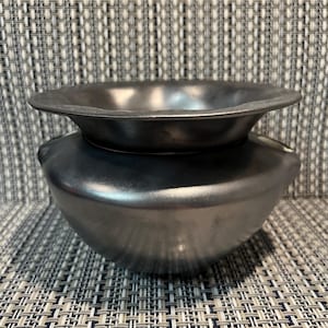 May include: A metallic dark grey bowl with a smooth, rounded body and a wide, flared rim. The surface has a subtle sheen, reflecting light across its curved exterior. The bowl features a distinct transition between the deep basin and the rim.