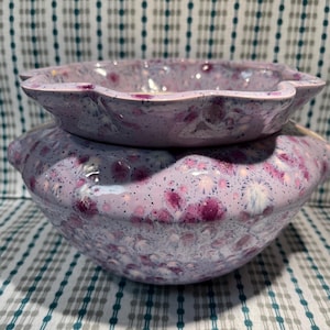 May include: Two-piece ceramic bowl set. The bowls are a light purple with a speckled pattern of pink and white. The top bowl is smaller and sits inside the larger bowl. The rims have a scalloped edge.