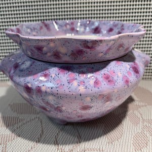 Peut inclure: Ensemble de deux bols en céramique. Les bols sont lavande avec une finition mouchetée et des motifs floraux roses. Le bol supérieur est plus petit et à bord festonné, le bol inférieur est plus grand et rond.