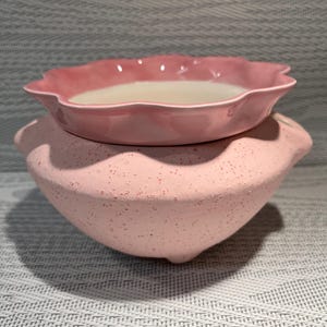 May include: A two-piece pink ceramic set. The top piece is a small, scalloped-edge bowl with a white liquid inside. The bottom piece is a larger, speckled pink bowl with a rounded shape. The set is designed for decorative use.