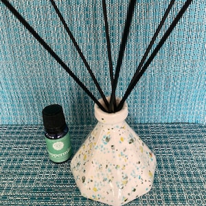 May include: A white ceramic diffuser bottle with a speckled design in blue, green, and yellow. The bottle is filled with black diffuser sticks and a small bottle of peppermint essential oil is in the foreground.