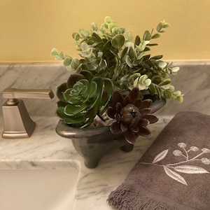 May include: A small black metal bathtub planter with a variety of green and brown succulents and greenery. The planter is sitting on a white marble countertop next to a white porcelain sink and a grey towel with white embroidery.