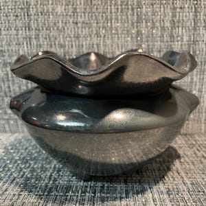 May include: A metallic charcoal grey ceramic bowl with a wide, ruffled, scalloped rim. The bowl features a smooth, reflective, and glossy finish that catches the light, creating subtle highlights across its curved, rounded body and edges.