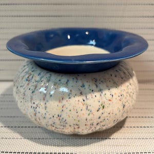 May include: A ceramic wax warmer featuring a round, white base with a multi-coloured speckled pattern and a ribbed texture. The top section is a wide, smooth, deep blue bowl with a curved rim, designed to hold wax melts for heating and fragrance.