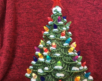 May include: A ceramic Christmas tree with a green glaze, adorned with colorful lights and a red star topper. The tree has a snow-like effect on the branches and sits on a green base. A white power cord extends from the base.