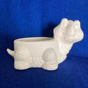May include: A white ceramic turtle planter with a smiling face, open top, and detailed shell design. The turtle has four legs, a tail, and a head with two eyes. The planter is designed to hold small plants or flowers.