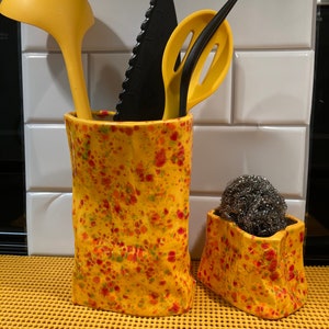 May include: Two yellow ceramic containers with a speckled pattern. The containers are filled with kitchen utensils, including a yellow ladle, a black knife, and a yellow slotted spoon. A silver scouring pad is in the smaller container.