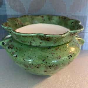 May include: A two-piece ceramic container with a light green speckled finish. The top piece has a scalloped edge and a white interior. The bottom piece is round with small handles on each side. The container is suitable for holding small items.
