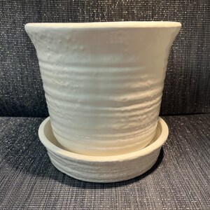 May include: A white ceramic plant pot with a textured, ridged design. The pot sits on a matching saucer. The pot has a flared rim and a cylindrical body. Suitable for indoor or outdoor use.
