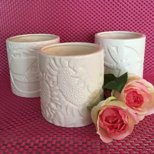 Peut inclure: Trois pots en céramique blanche avec des motifs en relief. Le pot de gauche a un motif géométrique, le pot du milieu a un motif de tournesol et le pot de droite a un motif de libellule. Les pots sont posés sur un fond rose avec des roses roses au premier plan.
