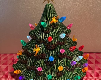 May include: A ceramic Christmas tree in a deep green glaze, adorned with colorful lights and small gold-colored ornaments. A gold glitter star tops the tree. The tree sits on a red and white patterned surface.