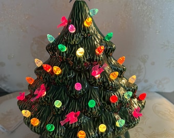 May include: A ceramic Christmas tree in a deep green glaze, adorned with colorful lights and small red bird ornaments. A dark blue star tops the tree. The tree is lit with various colored lights, including orange, green, pink, and yellow.