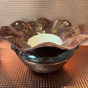 May include: A ceramic bowl featuring a deep brown exterior with a glossy finish and a wide, ruffled, wavy rim. The interior of the bowl is a smooth, solid off-white colour. The bowl has a rounded base and small side handles, showcasing a dark, mottled glaze texture throughout the outer surface.