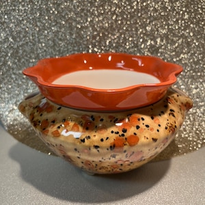 May include: A two-piece ceramic candle holder. The top piece is a bright orange, scalloped-edge bowl filled with white wax. The bottom piece is a larger, tan-coloured bowl with orange and black speckled designs.